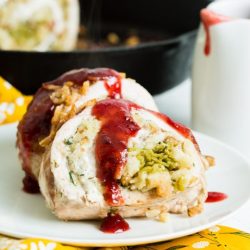 Rolled turkey with stuffing and cranberry sauce on a white plate, with a skillet and sauce cup in the background.