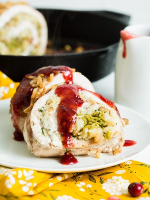 Rolled turkey with stuffing and cranberry sauce on a white plate, with a skillet and sauce cup in the background.