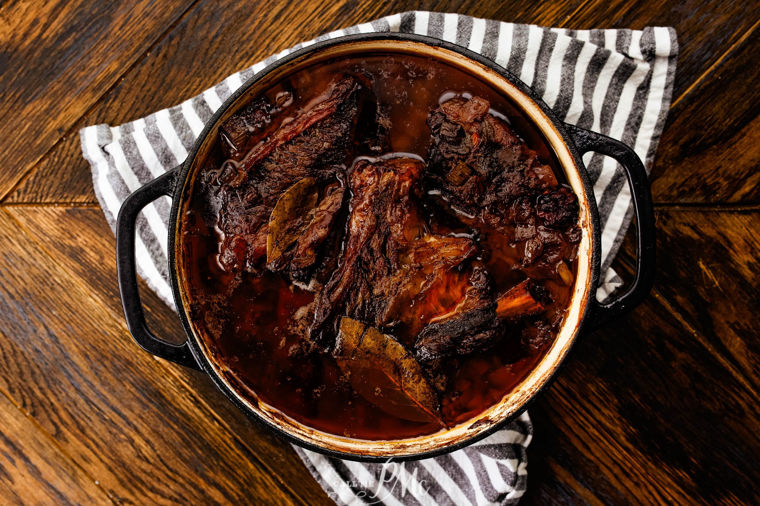 A pot of braised beef with bay leaves, in rich brown sauce, on a striped cloth.