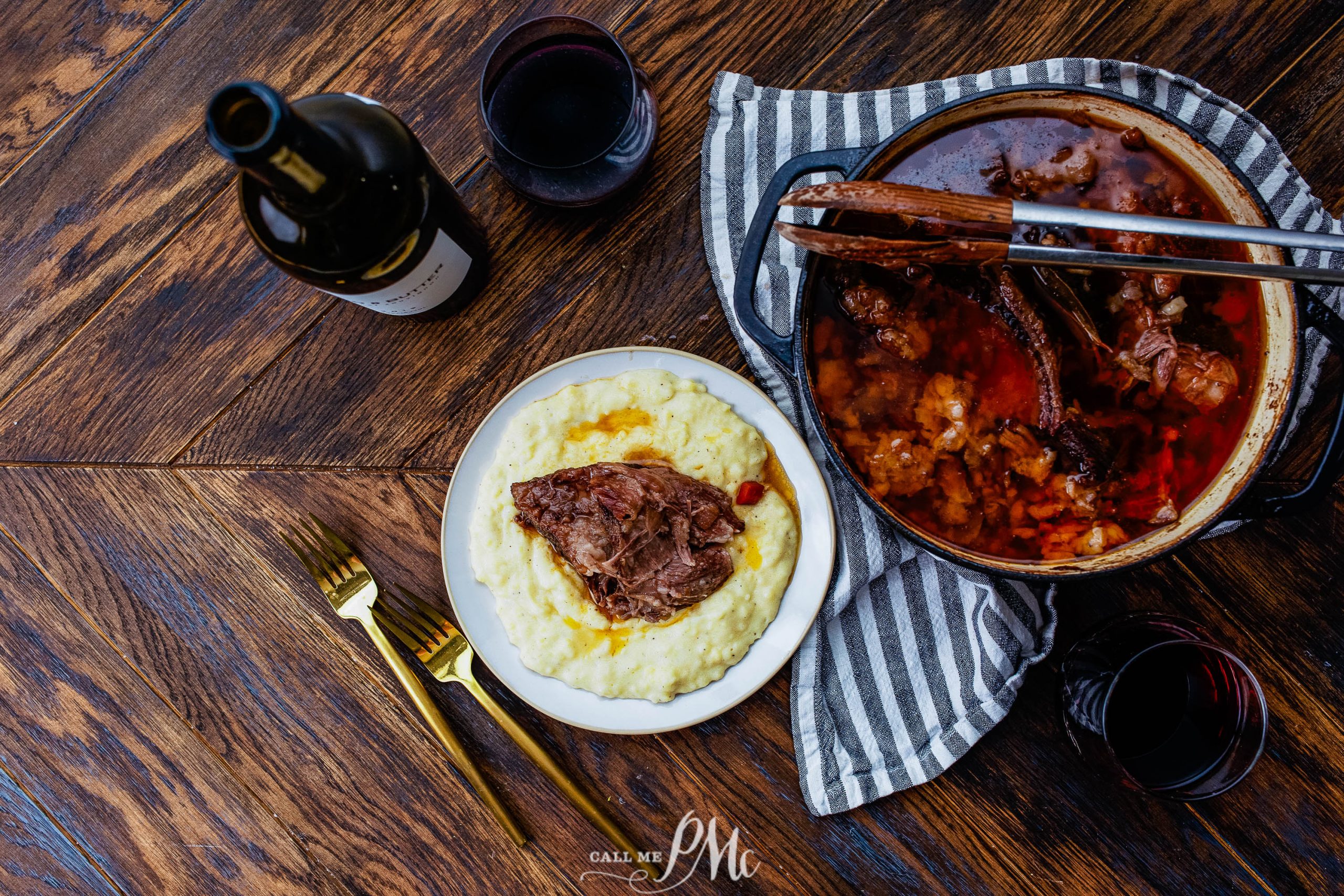 Plate of mashed potatoes topped with meat beside a pot of stew, wine bottle, and glass on a wooden table with utensils.