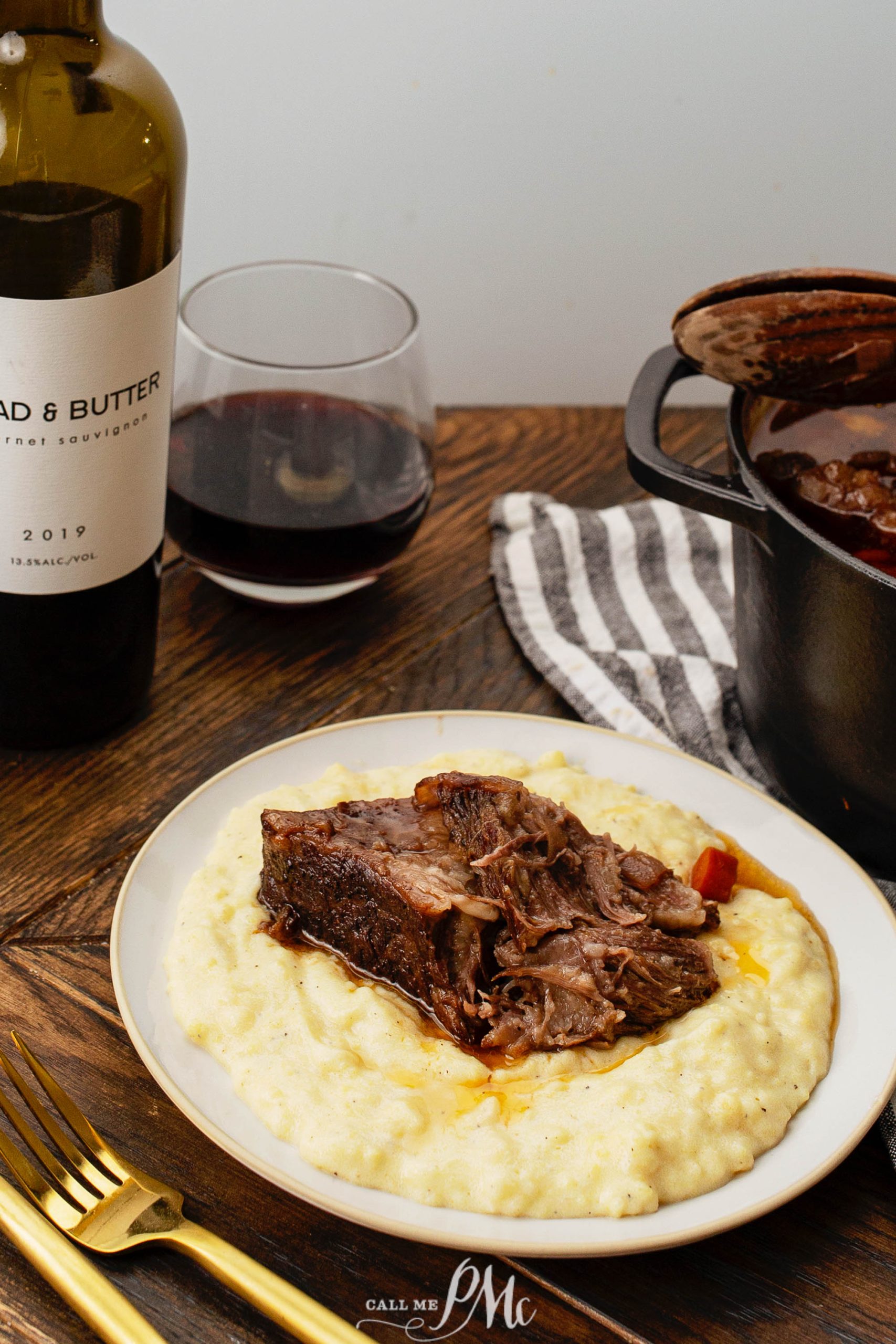 A plate with braised beef on creamy polenta, beside a glass of red wine and a bottle, with a striped napkin and fork. A pot of beef stew is visible in the background.