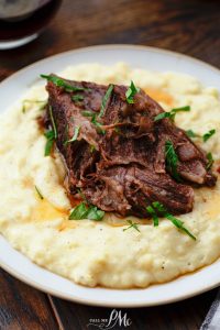 A serving of tender pot roast sits atop creamy mashed potatoes, garnished with fresh herbs, on a white plate.