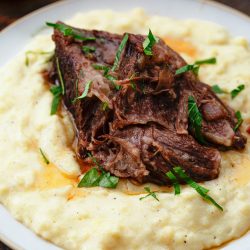 A serving of tender pot roast sits atop creamy mashed potatoes, garnished with fresh herbs, on a white plate.