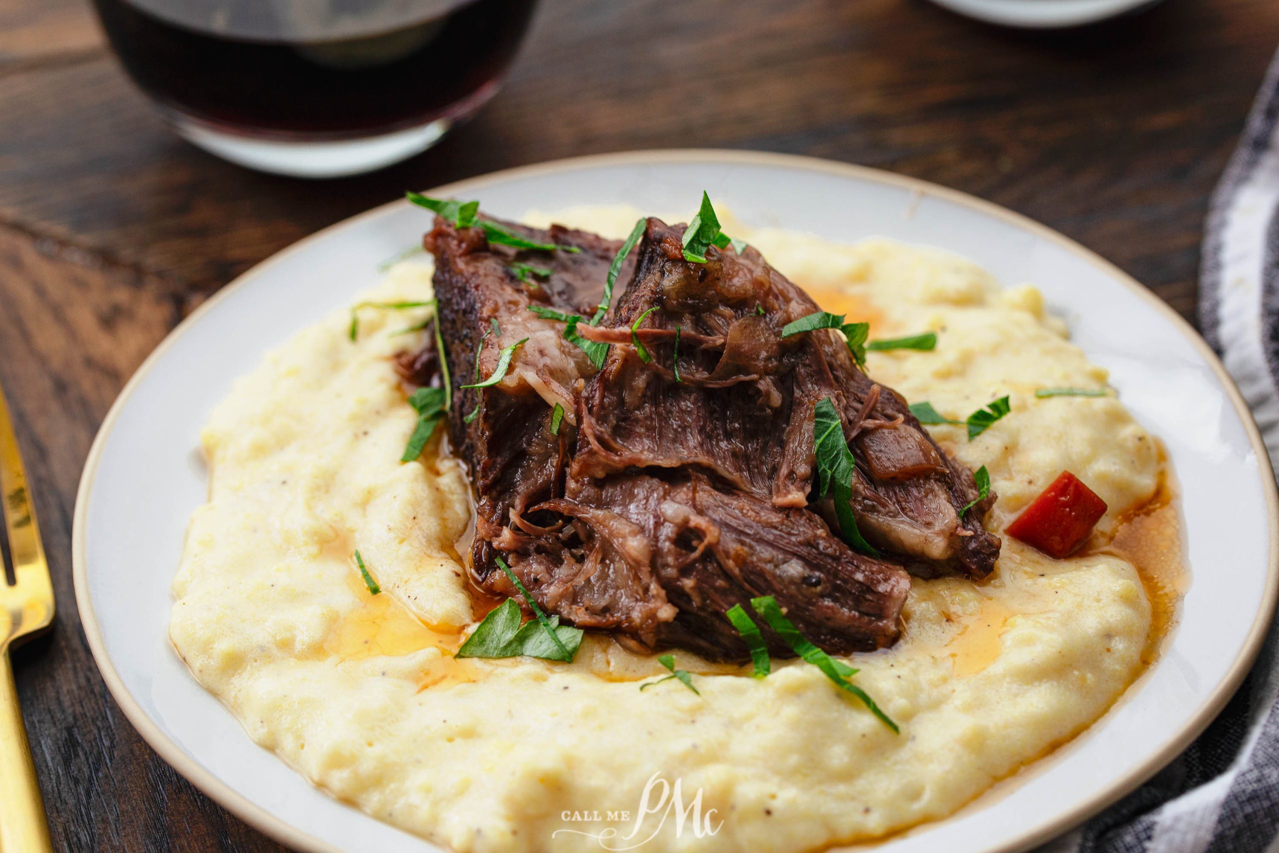 A plate of shredded braised beef on creamy mashed potatoes, garnished with herbs, served on a wooden table next to a glass of red wine.