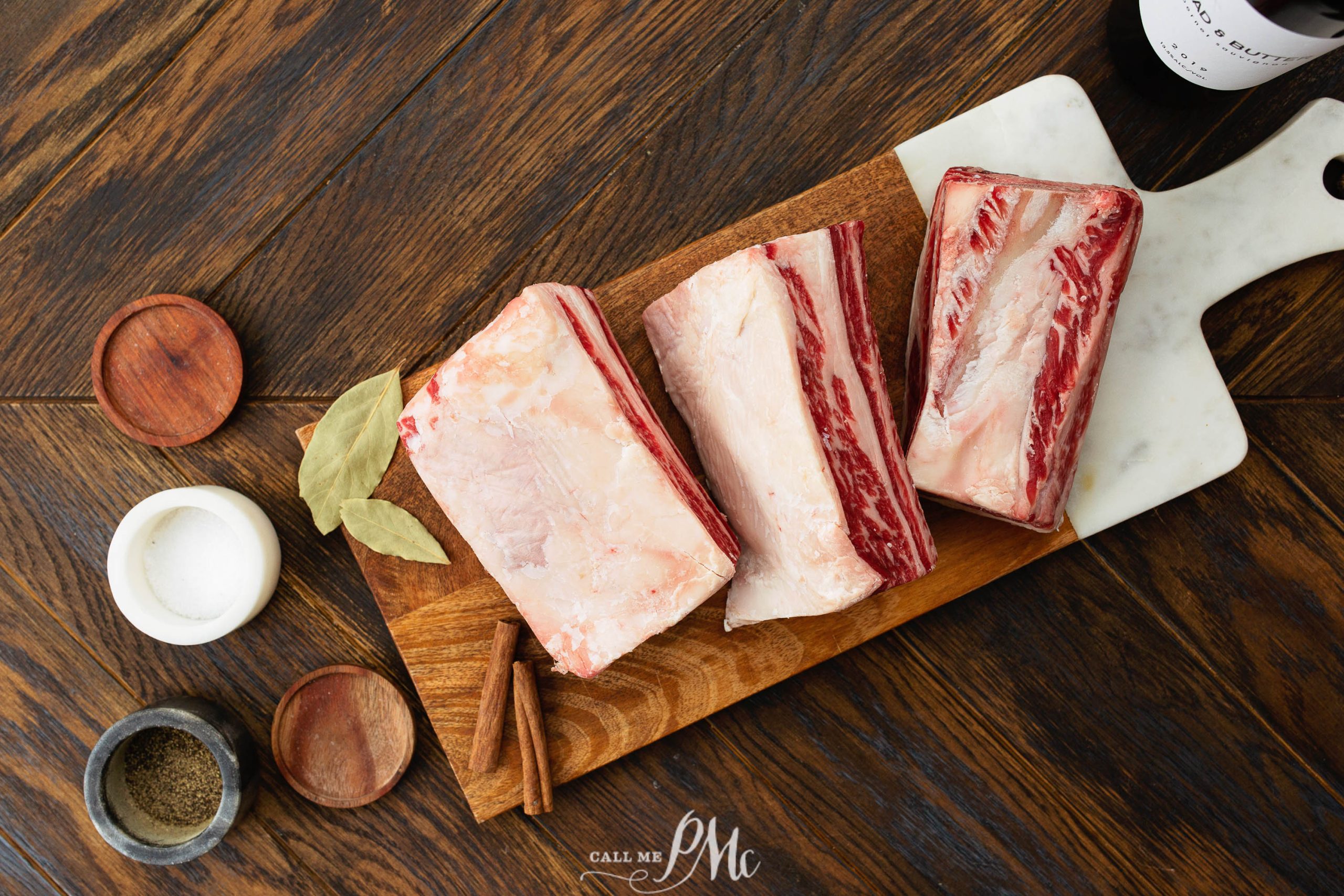 Three raw beef short ribs on a wooden cutting board, accompanied by bay leaves, cinnamon sticks, a small bowl of salt, and a small bowl of pepper, placed on a wooden table.