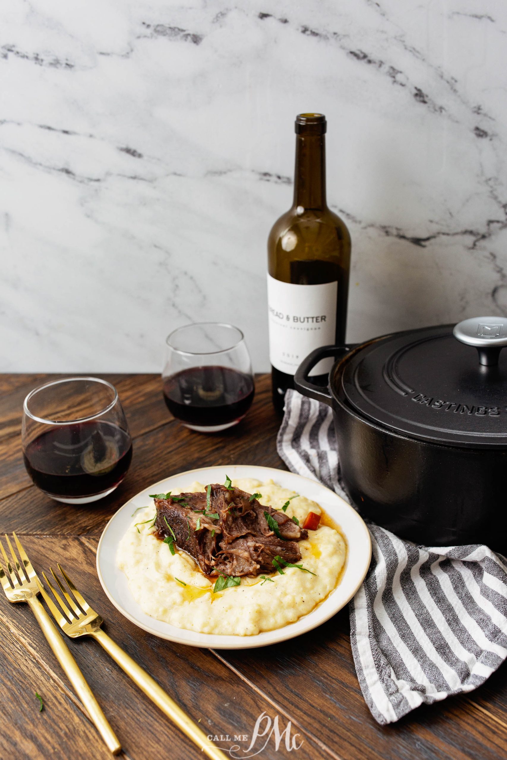 A plate of creamy polenta topped with braised short ribs, beside a bottle of red wine, two wine glasses, a black pot, and gold cutlery on a wooden table with a striped cloth.