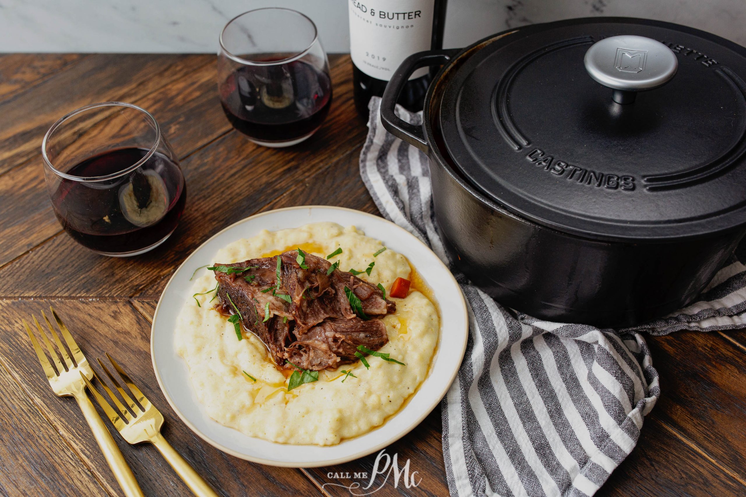 Braised beef served on a bed of creamy polenta, with two glasses of red wine, a black Dutch oven, and gold utensils on a wooden table.