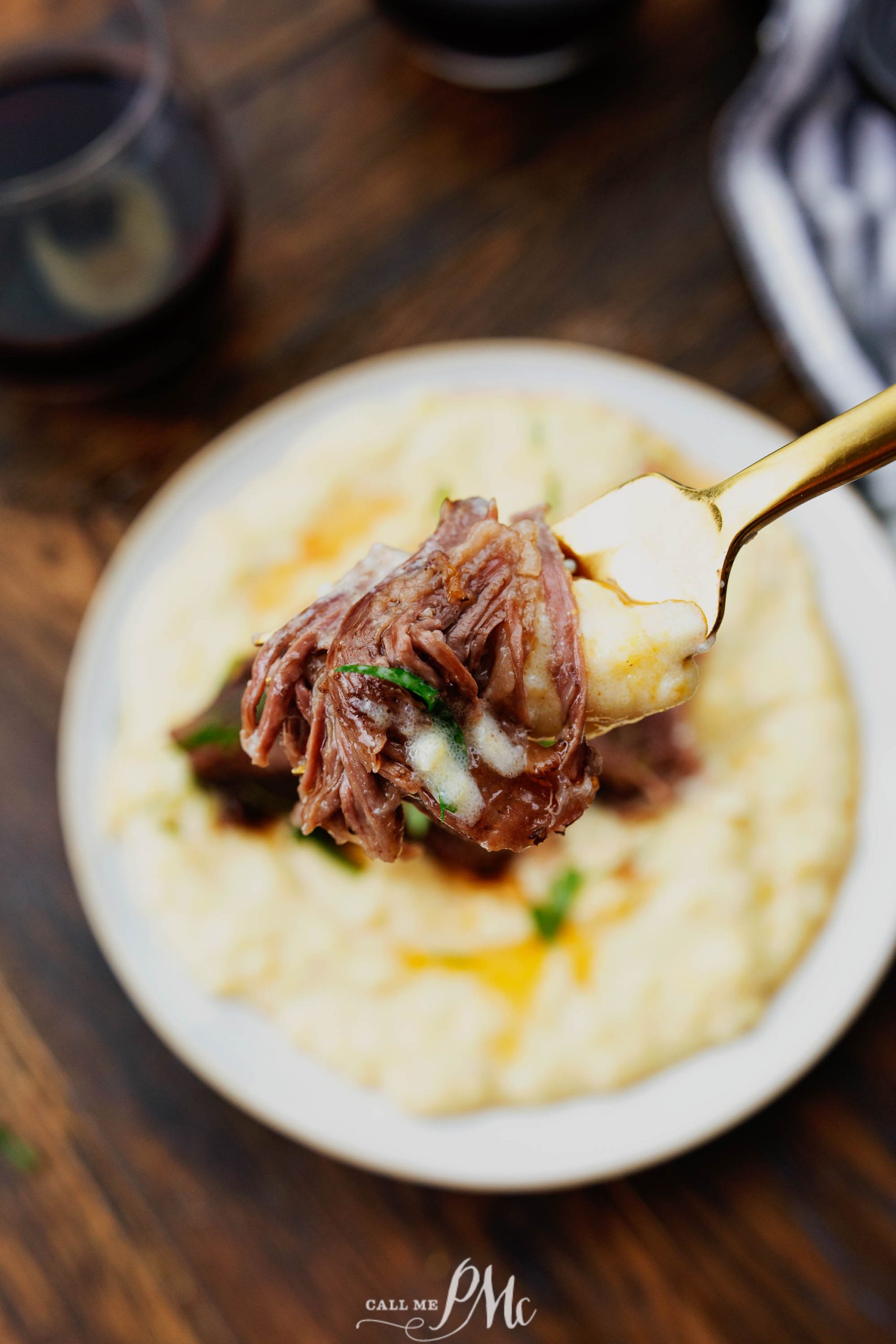 A fork holding shredded beef over a plate of creamy mashed potatoes on a wooden table, with a glass of red wine in the background.