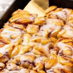 A tray of freshly baked cinnamon rolls topped with a layer of white icing, with a gold spatula resting on the side.