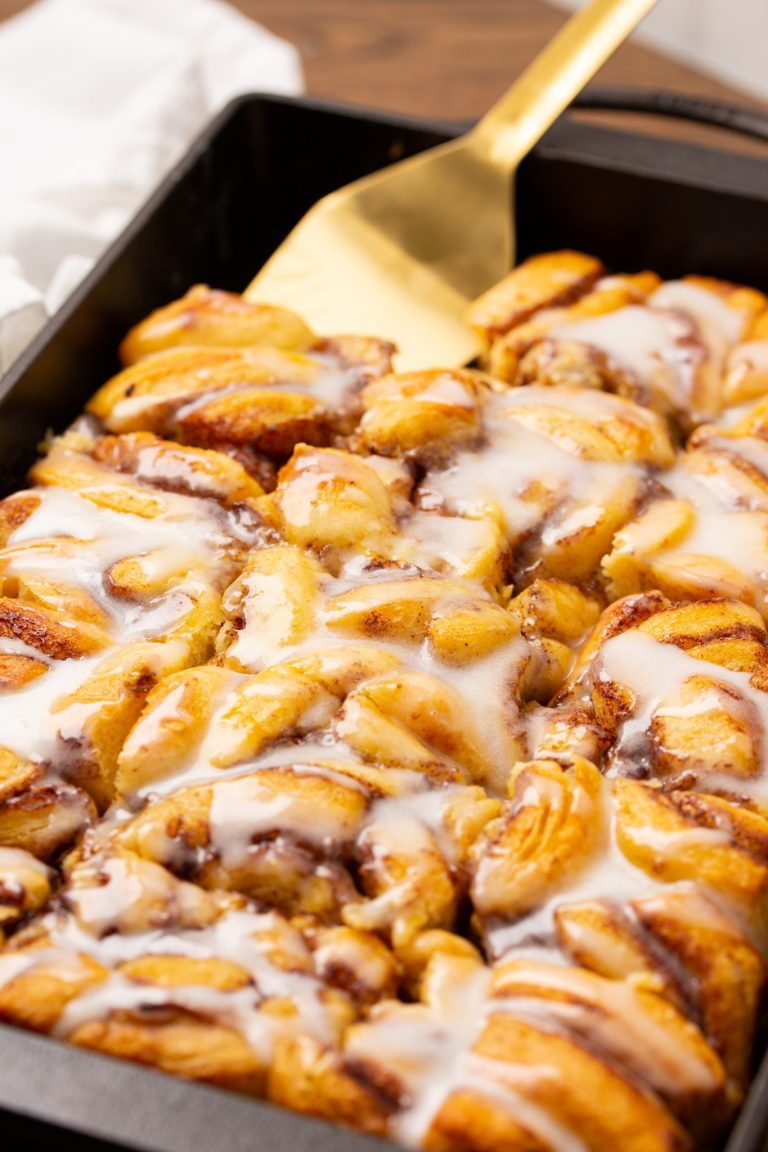 A tray of freshly baked cinnamon rolls topped with a layer of white icing, with a gold spatula resting on the side.