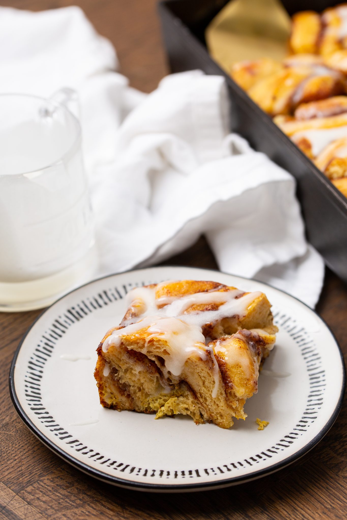 A cinnamon roll with icing on top sits on a plate, next to a jug of milk and a pan of rolls, with a white cloth nearby.