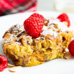 A slice of baked French toast topped with raspberries and powdered sugar on a white plate.