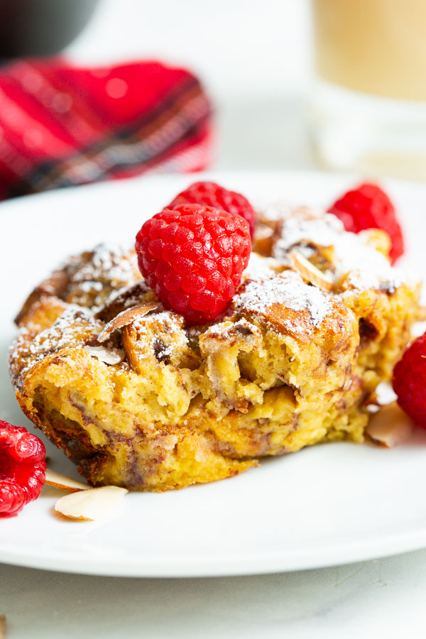 A slice of baked French toast topped with raspberries and powdered sugar on a white plate.