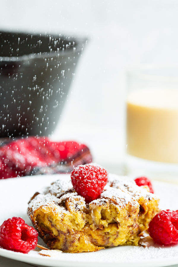 Bread pudding topped with powdered sugar and raspberries on a white plate, with a drink and black dish in the background.