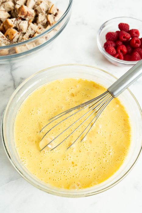 A bowl of egg mixture with a whisk, a bowl of cubed bread, and a bowl of raspberries on a marble countertop.