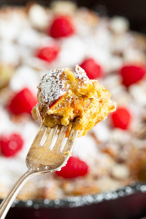 Close-up of a fork holding a piece of French toast dusted with powdered sugar. Blurred background shows more French toast and several raspberries.