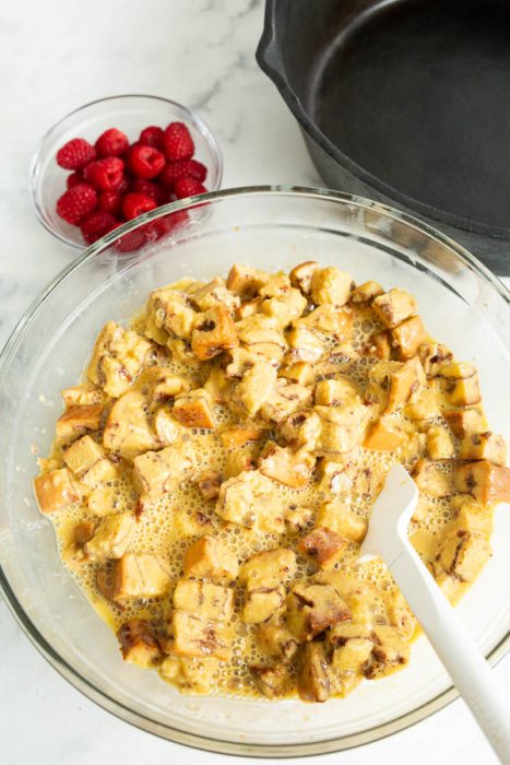 A clear bowl of bread cubes soaked in egg mixture with a spatula, next to a bowl of raspberries on a marble surface. A cast iron skillet is in the background.