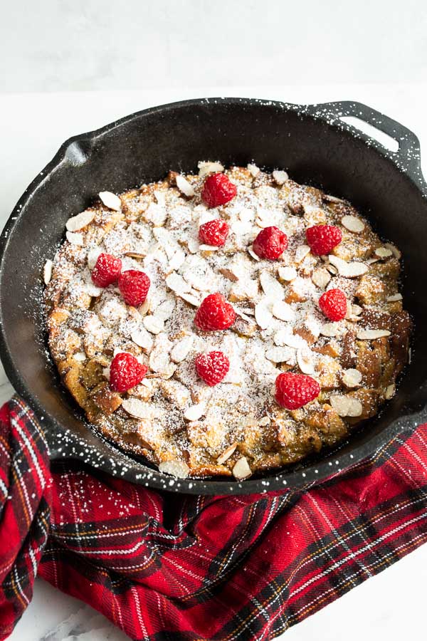 A cast iron skillet holds a baked dessert topped with powdered sugar, raspberries, and almond slices. A red plaid cloth is placed beside the skillet.