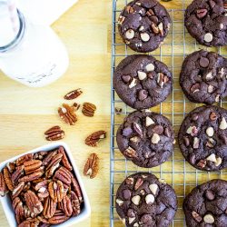 Chocolate cookies with pecans and white chocolate chips on a cooling rack, next to a bowl of pecans and a bottle of milk with a straw, set on a wooden surface.