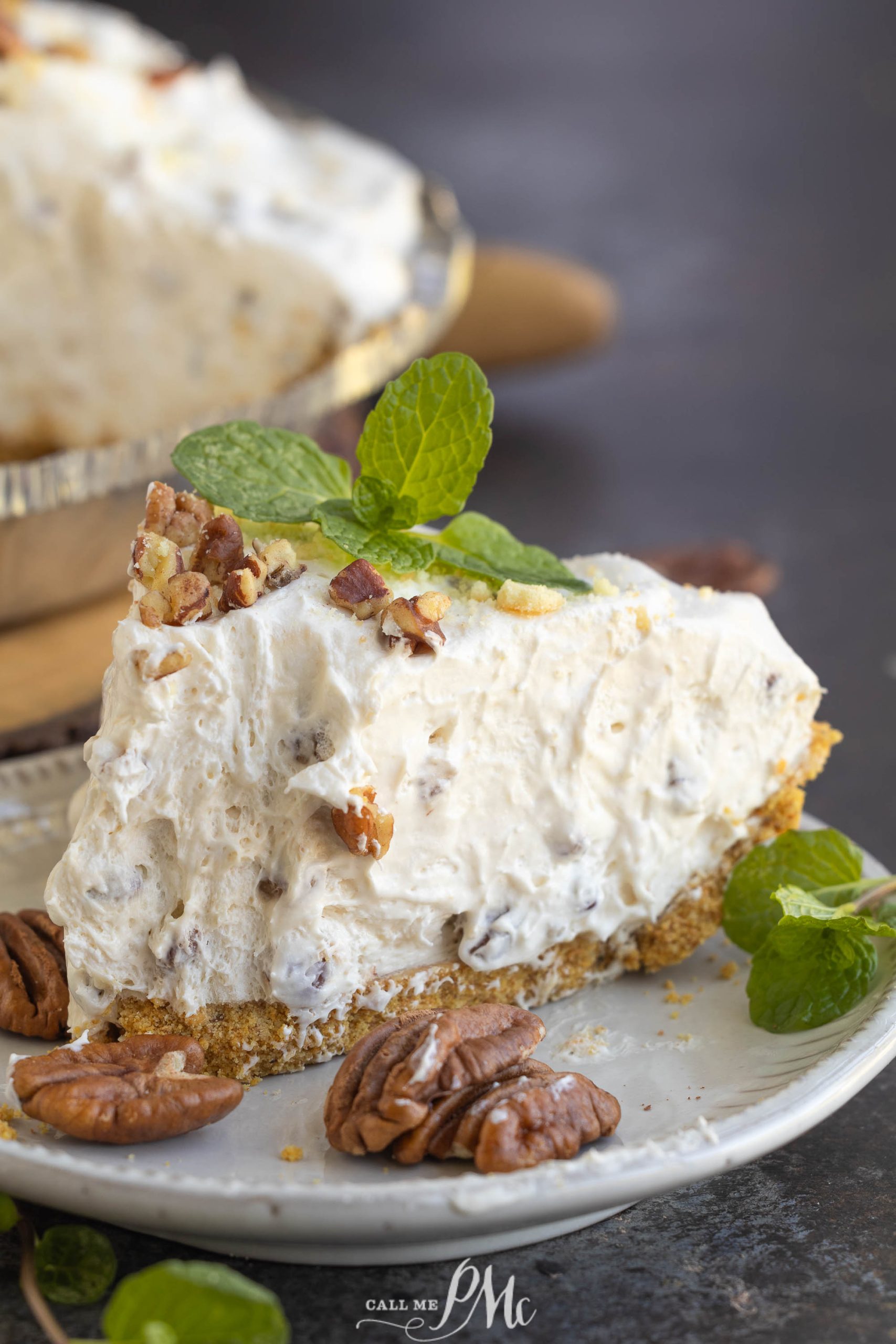 A slice of creamy, nut-topped pie on a plate with pecans and mint leaves garnish.