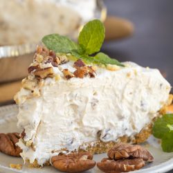 A slice of creamy cheesecake with a graham cracker crust, topped with pecans and garnished with mint leaves on a plate.