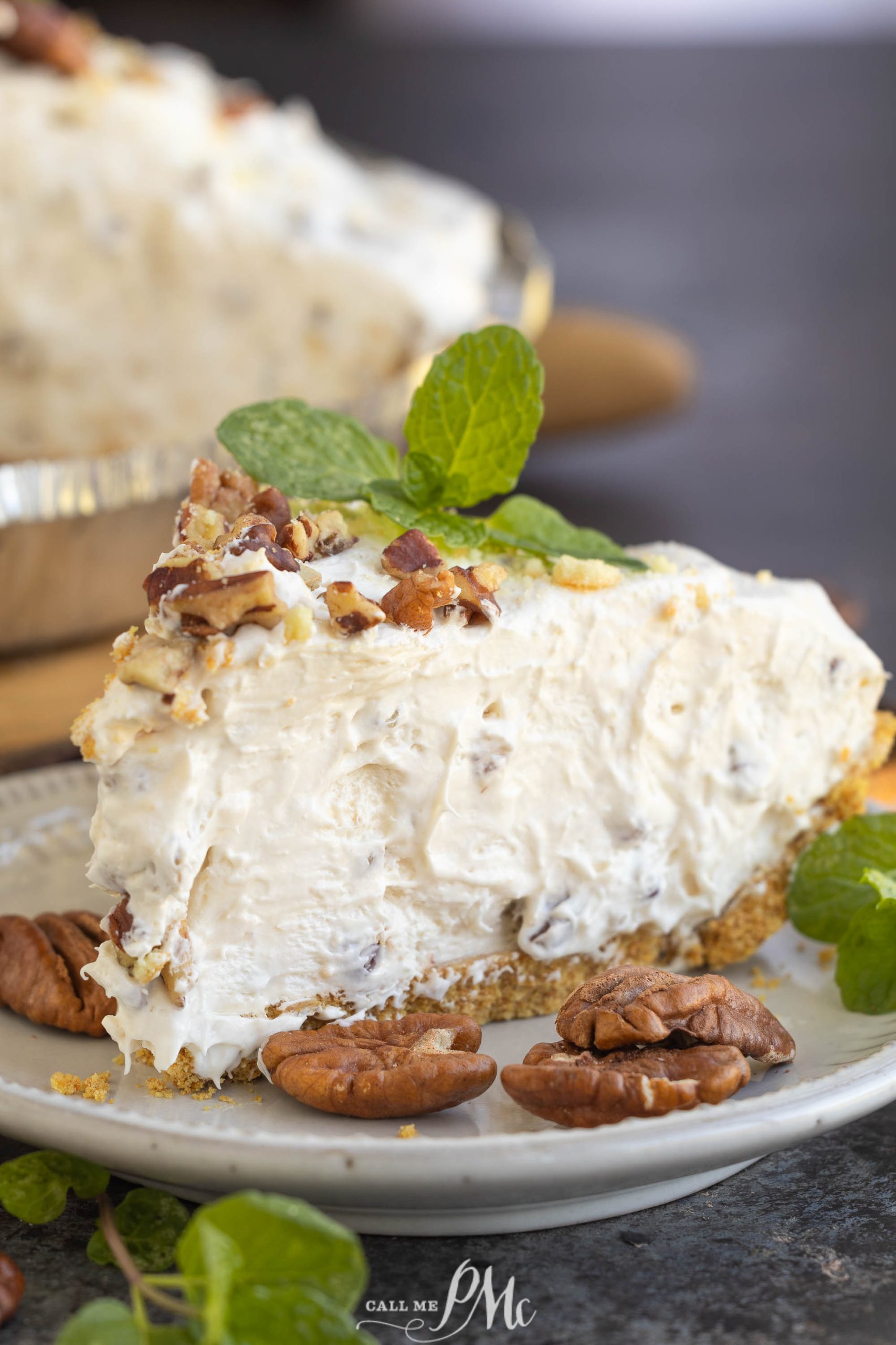 A slice of creamy cheesecake with a graham cracker crust, topped with pecans and garnished with mint leaves on a plate.