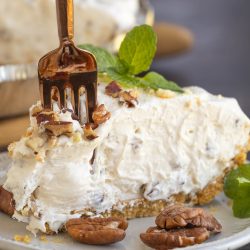 A fork slices into a creamy pie topped with chopped pecans, displayed on a plate with whole pecans and mint leaves.