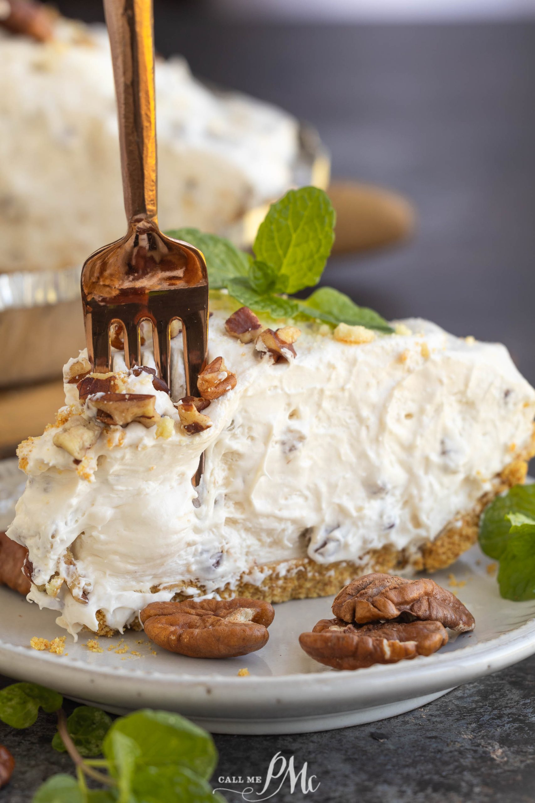 A fork slices into a creamy pie topped with chopped pecans, displayed on a plate with whole pecans and mint leaves.