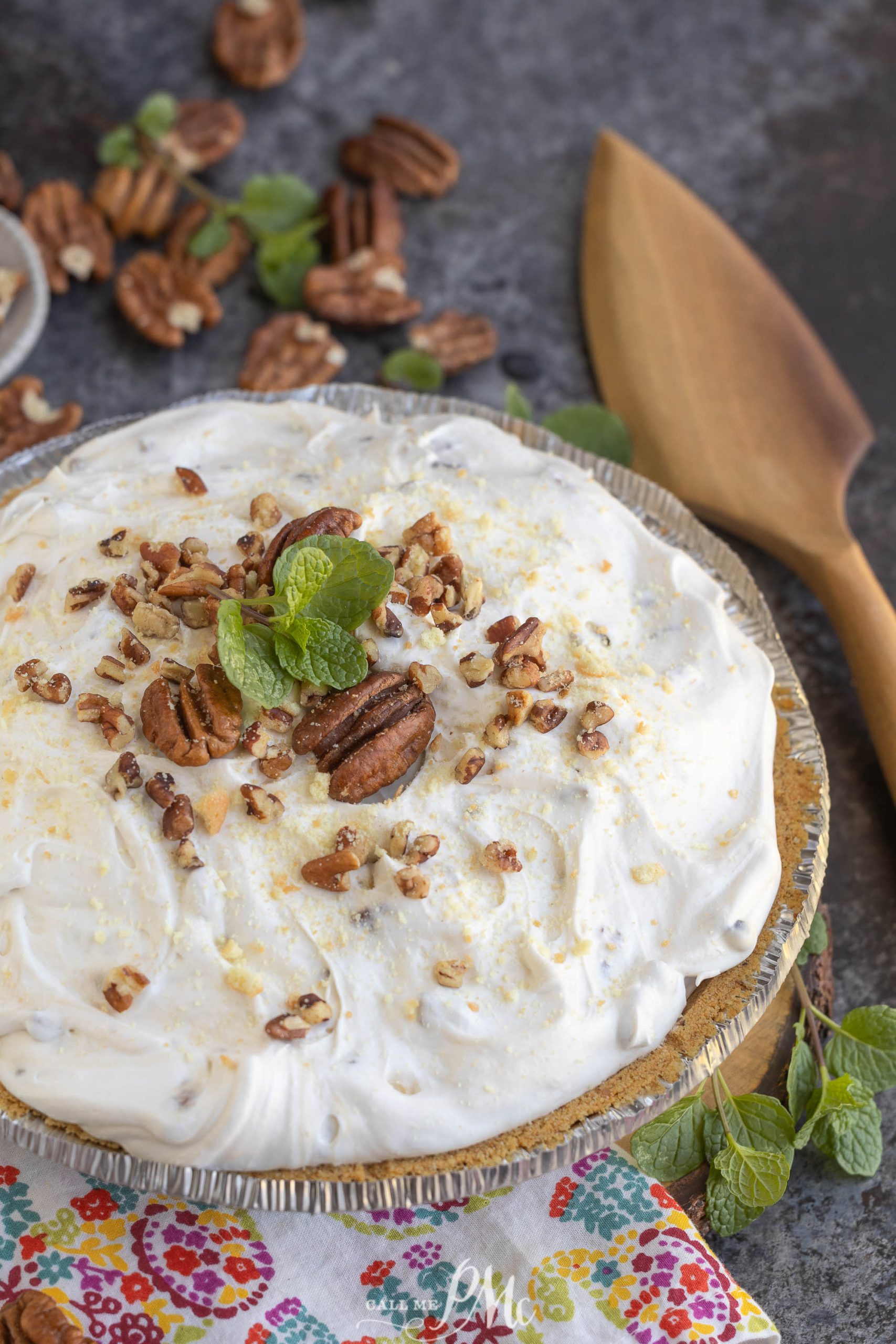 A creamy pie topped with chopped pecans and a mint garnish, surrounded by scattered pecans, a wooden spatula, and a colorful cloth.