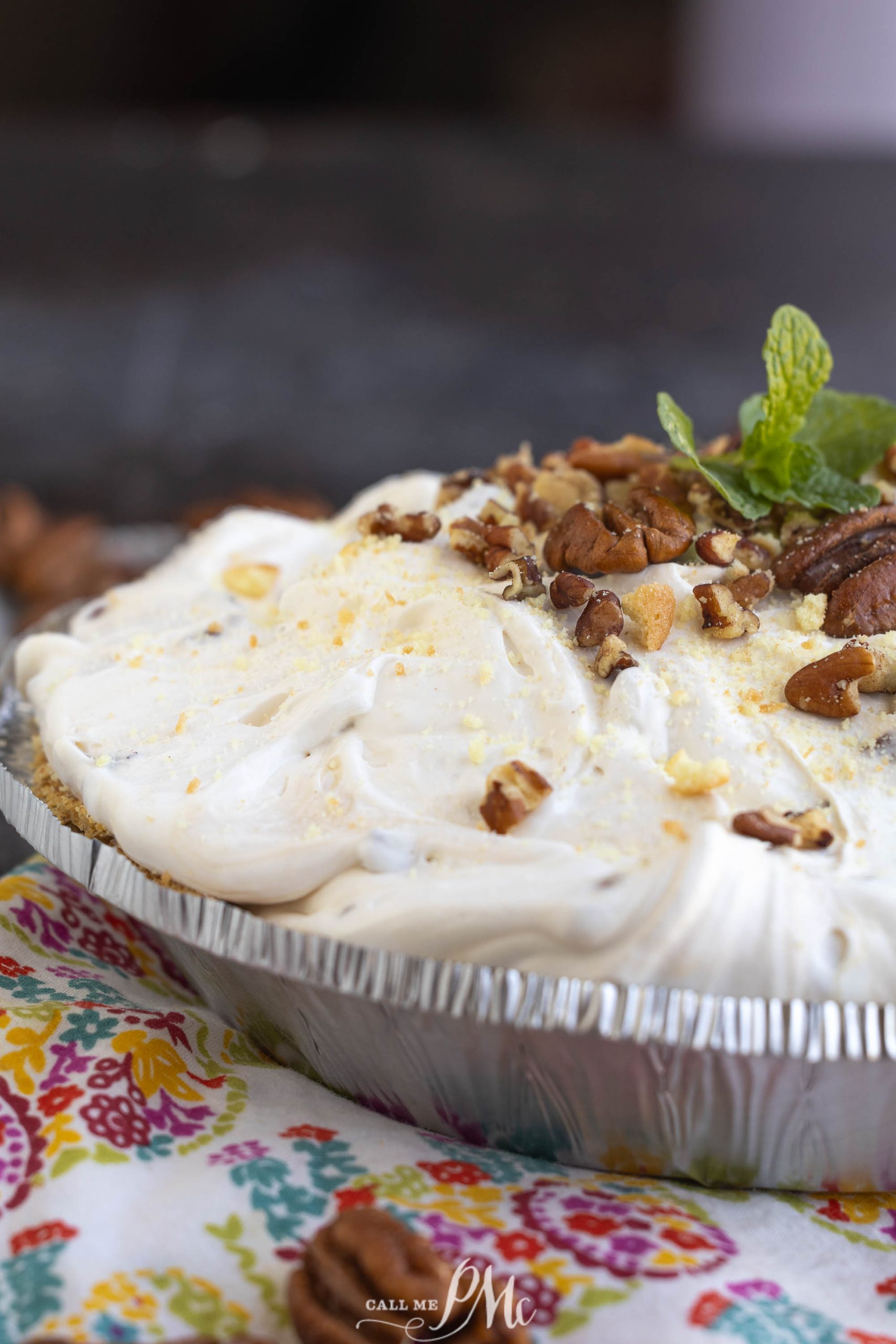 Cream pie topped with whipped cream, pecans, and a sprig of mint in a foil pie dish on a colorful floral cloth.