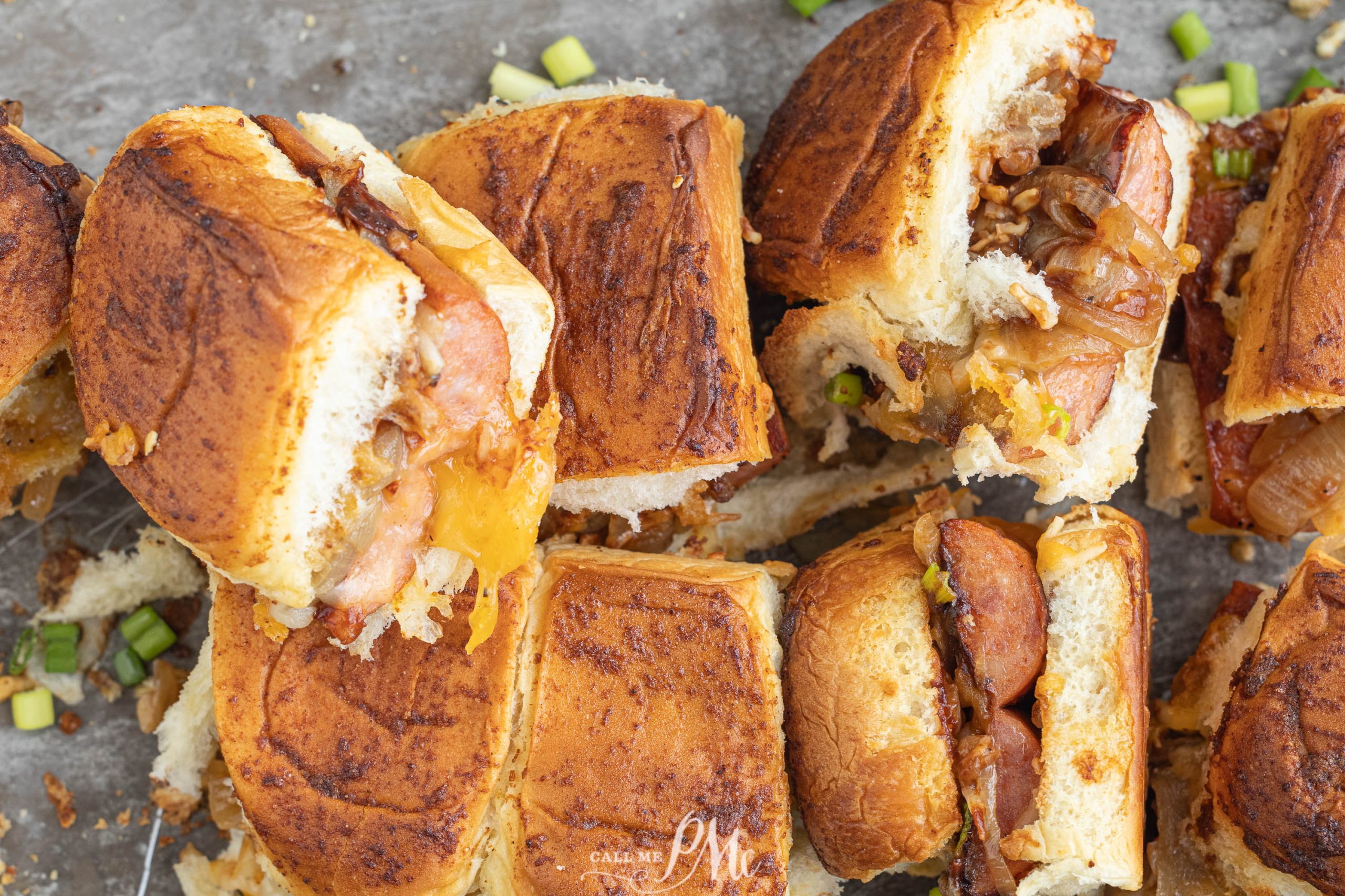 Close-up of sliced hot dog sliders with melted cheese, caramelized onions, and chopped green onions on toasted bread, arranged on a gray surface.