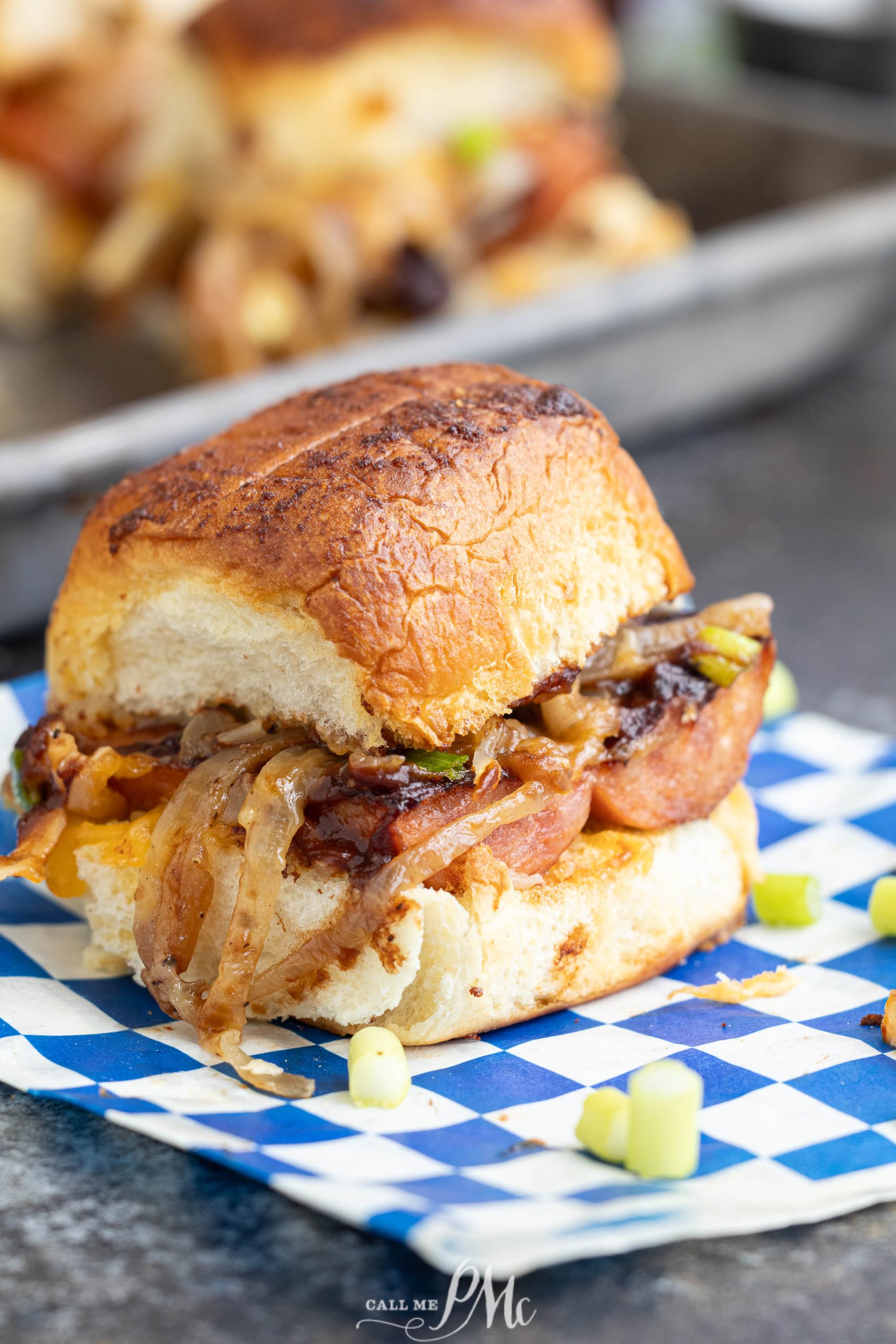 A slider with grilled sausage, caramelized onions, and green onions on a toasted bun, placed on a blue and white checkered paper.