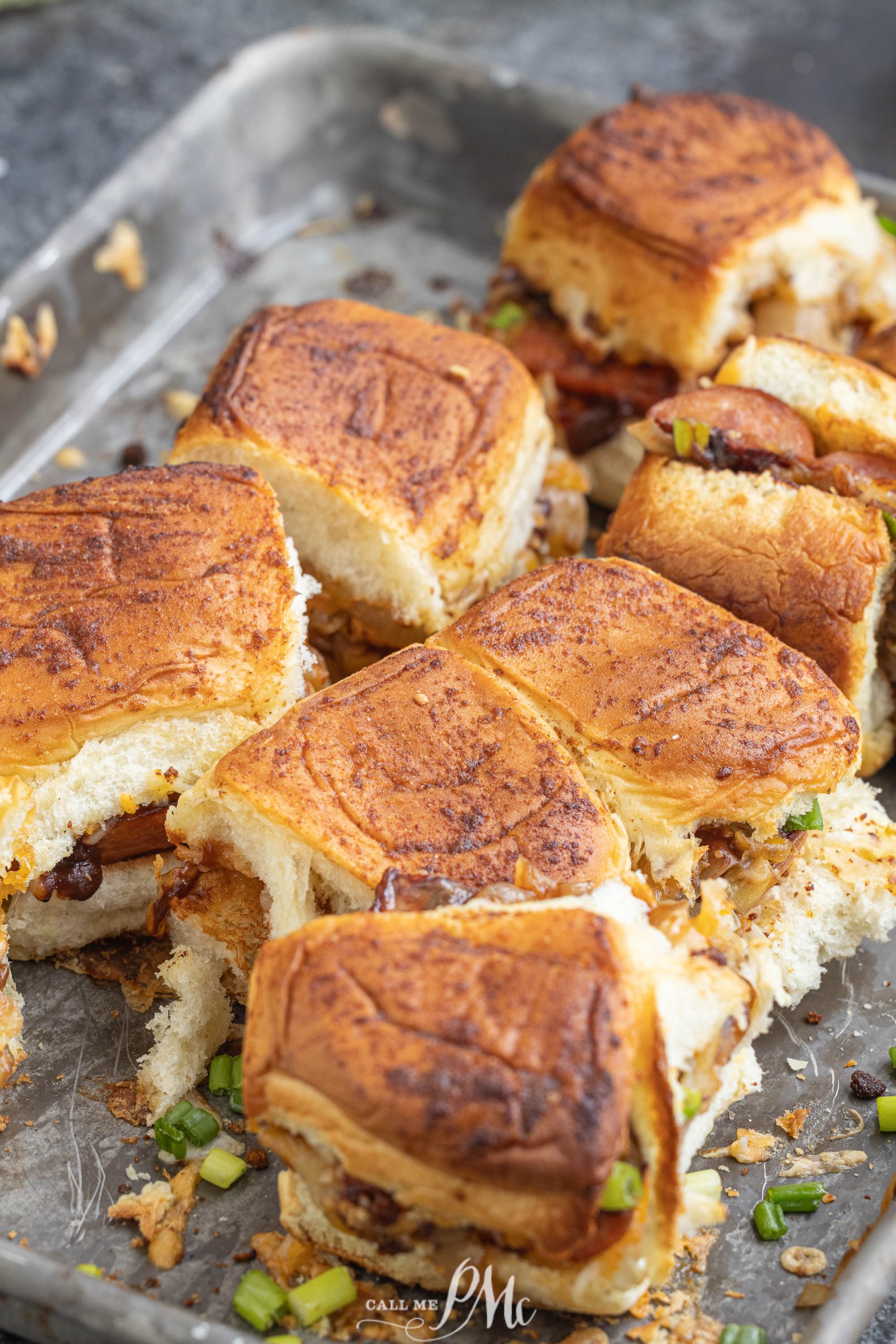 A tray of browned sliders filled with cooked meat and cheese, topped with chopped green onions.