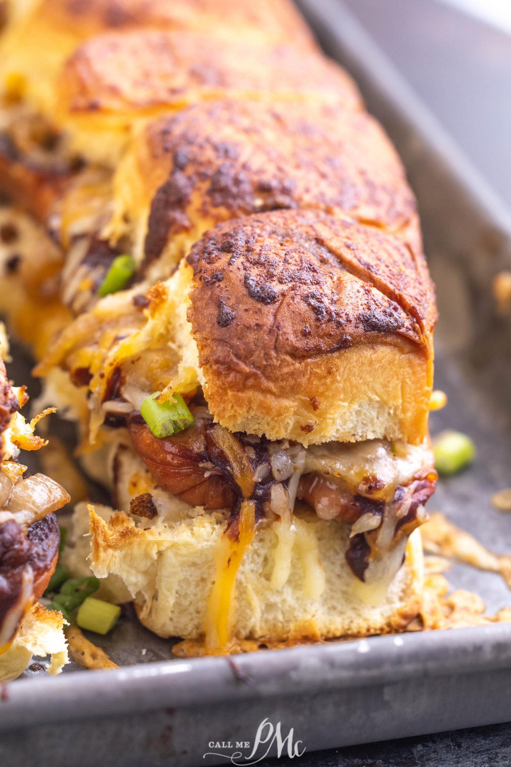 Close-up of baked sliders with melted cheese, onions, and green toppings on a baking tray.