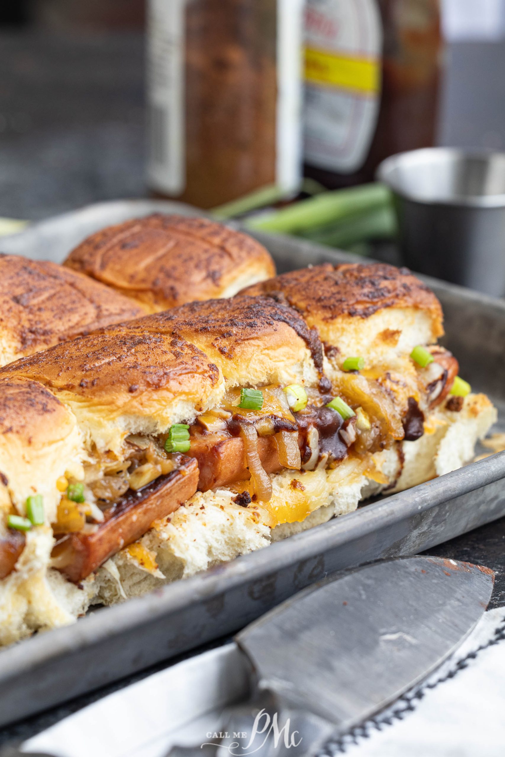 Toasted bread rolls filled with sausage, melted cheese, onions, and garnished with green onions, displayed on a tray with a knife nearby.