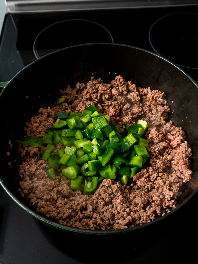 Ground beef and chopped green bell peppers cooking in a black skillet.