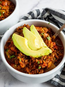 A bowl of chili topped with avocado slices, placed on a table with a striped napkin nearby.