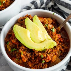 A bowl of chili topped with avocado slices, placed on a table with a striped napkin nearby.