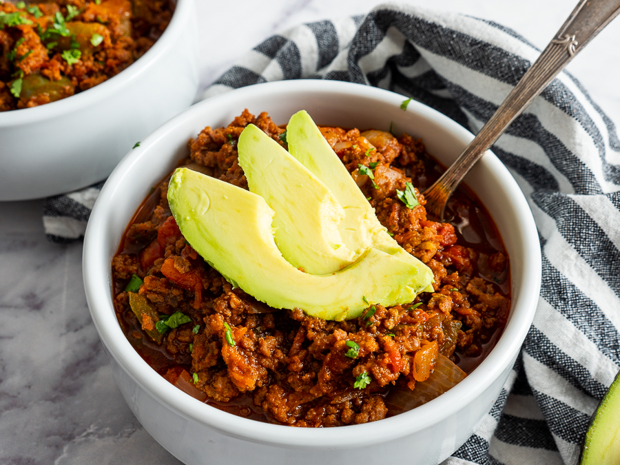 A bowl of chili topped with avocado slices, placed on a marble surface with a striped cloth.