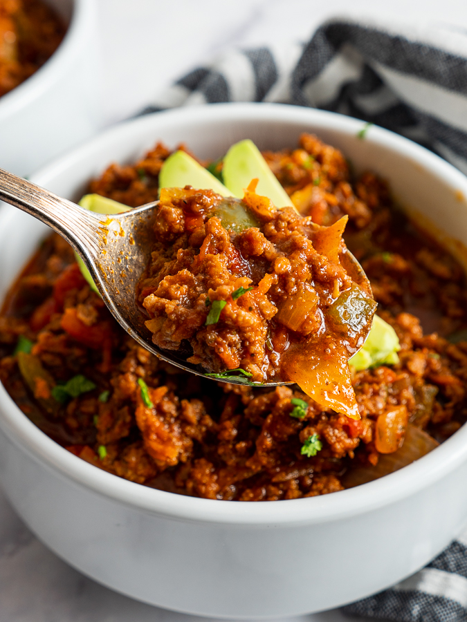 A spoonful of Spicy Chipotle Chiliwith ground meat, onions, and peppers held above a white bowl, garnished with avocado slices.