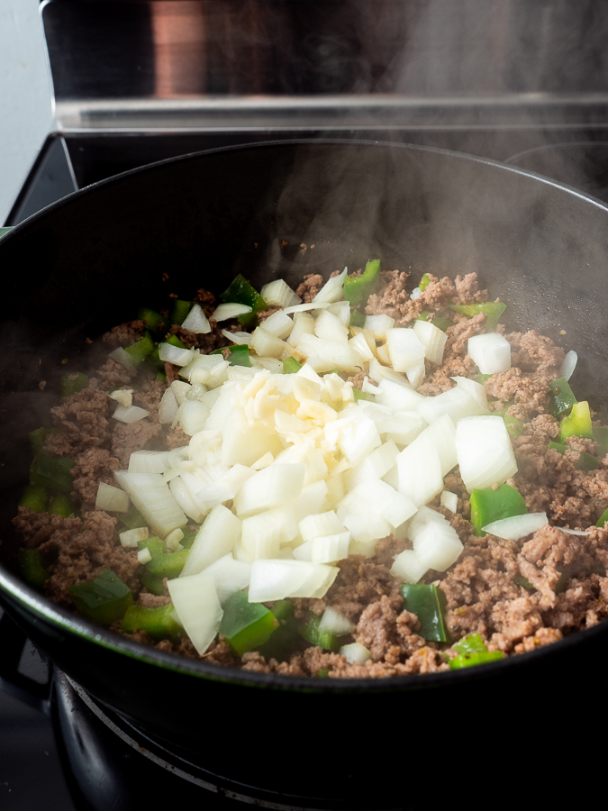 Ground beef, chopped onions, and green peppers cooking in a pan on a stovetop with visible steam.