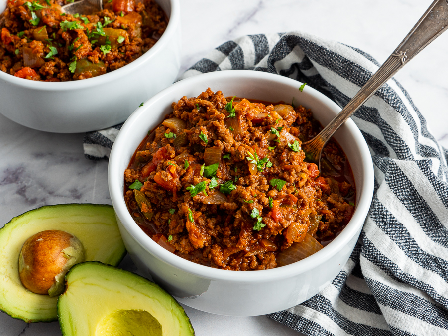 Two bowls of Spicy Chipotle Chili with a spoon, garnished with herbs. Half an avocado is on the side, and a striped cloth napkin is visible.