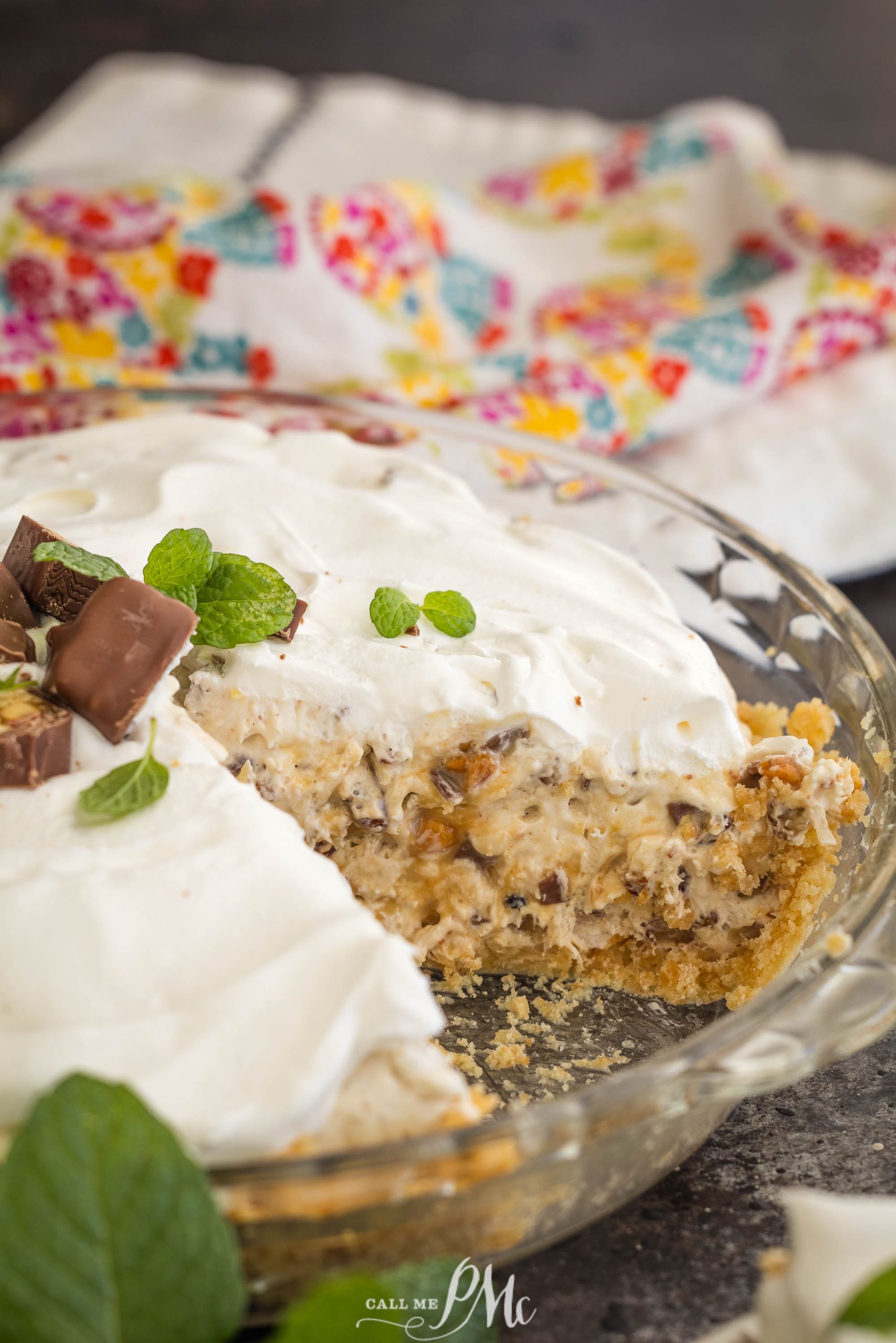 A creamy million dollar pie topped with whipped cream, mint leaves, and chocolate pieces, with a slice missing. A colorful cloth is in the background.