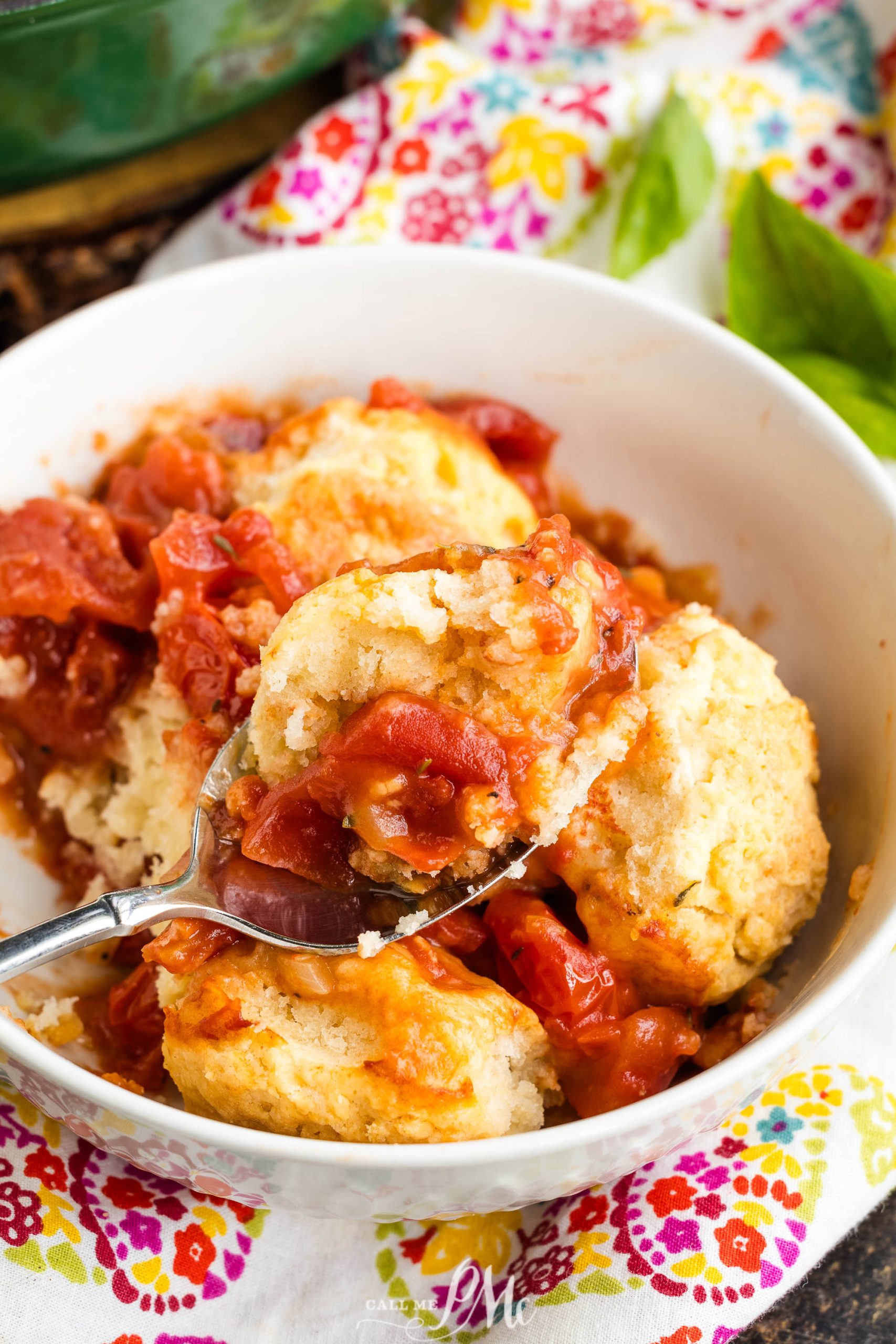 A bowl with tomato cobbler and biscuit topping, shown on a spoon, rests on a colorful floral fabric.