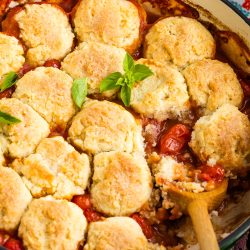 A casserole dish with golden-brown biscuits on top of a tomato-based filling, garnished with fresh basil, and a wooden spoon resting inside.