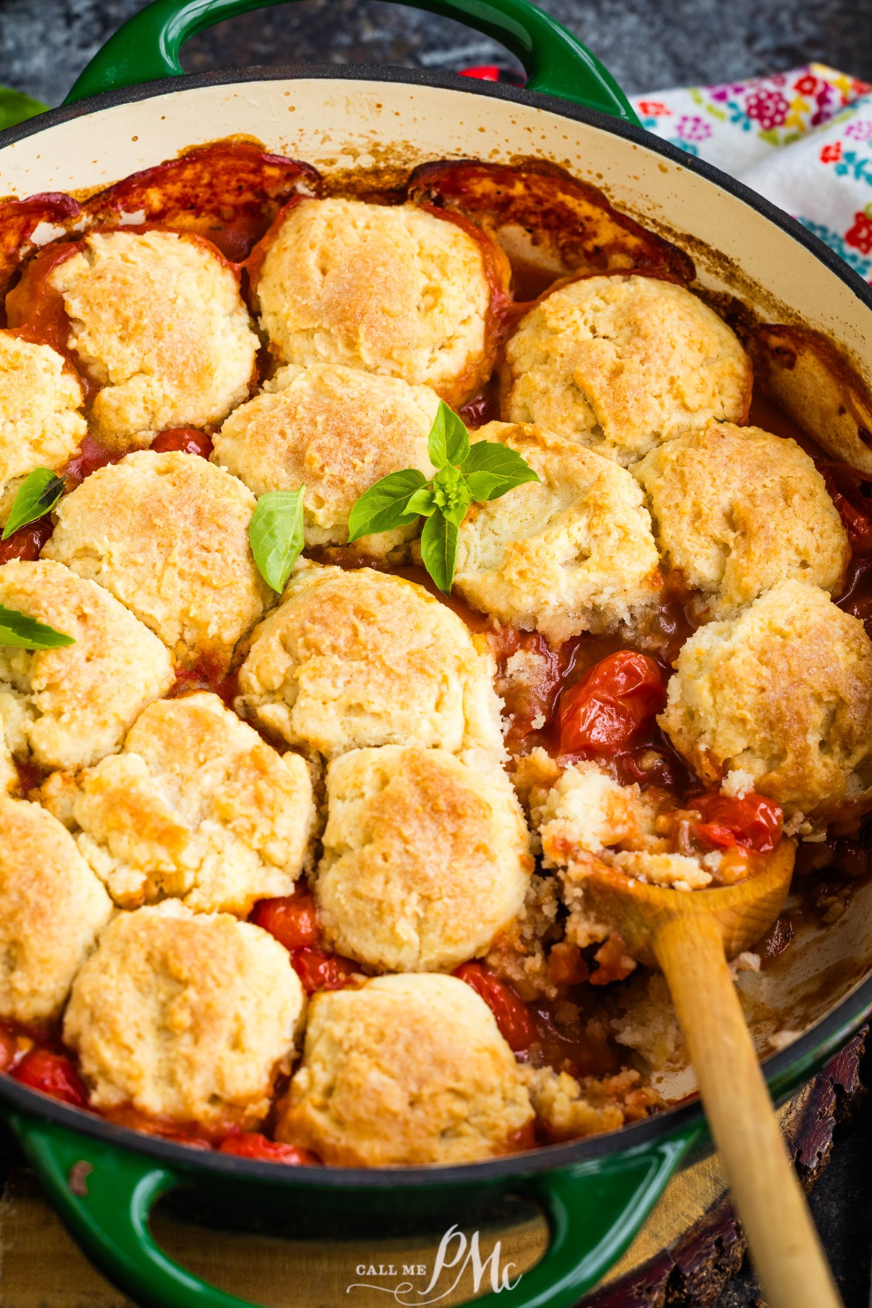 A casserole dish with golden-brown biscuits on top of a tomato-based filling, garnished with fresh basil, and a wooden spoon resting inside.