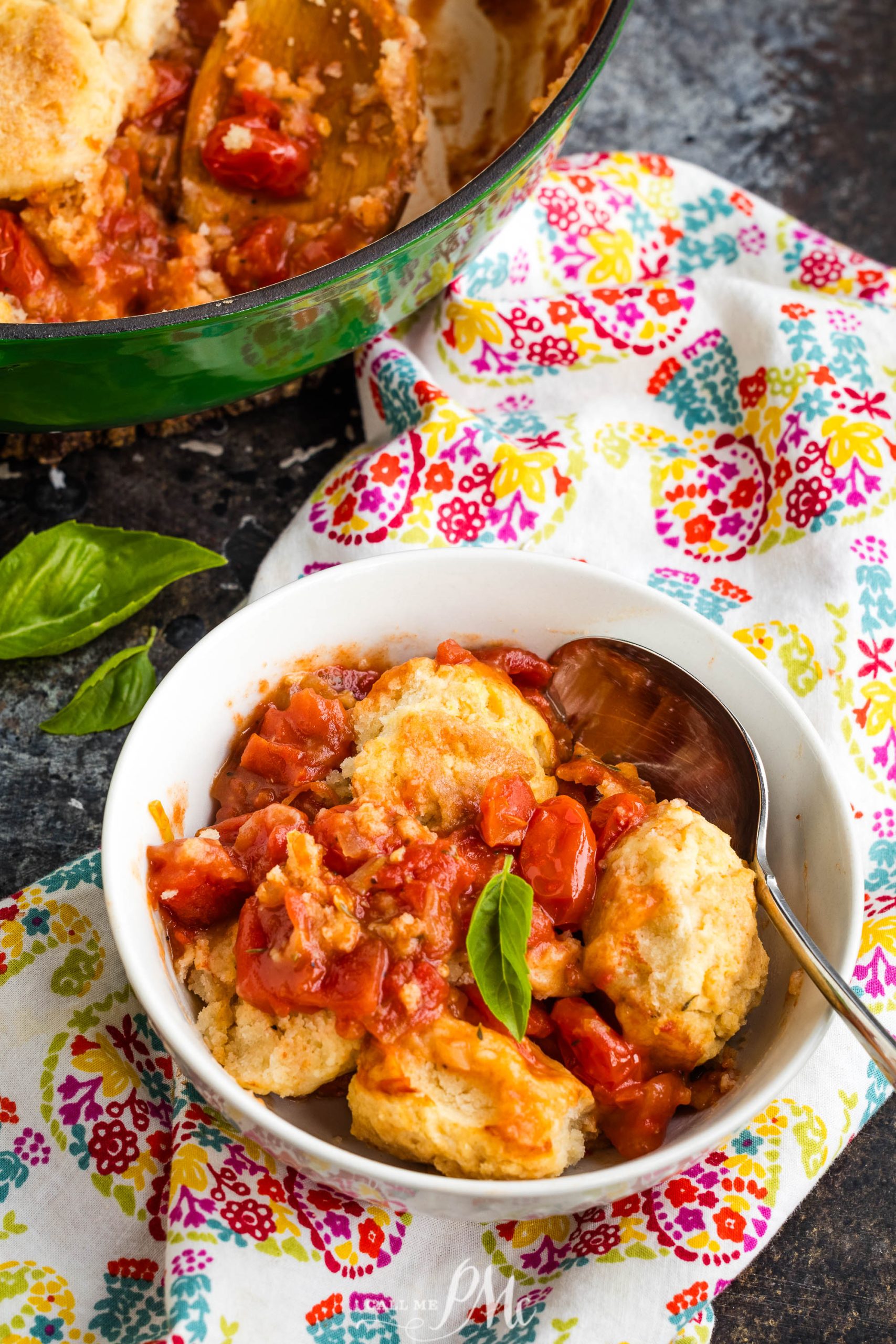 A bowl of tomato cobbler with a biscuit topping is placed on a colorful floral napkin. A basil leaf and a spoon rest in the bowl. A green pot with more cobbler is in the background.