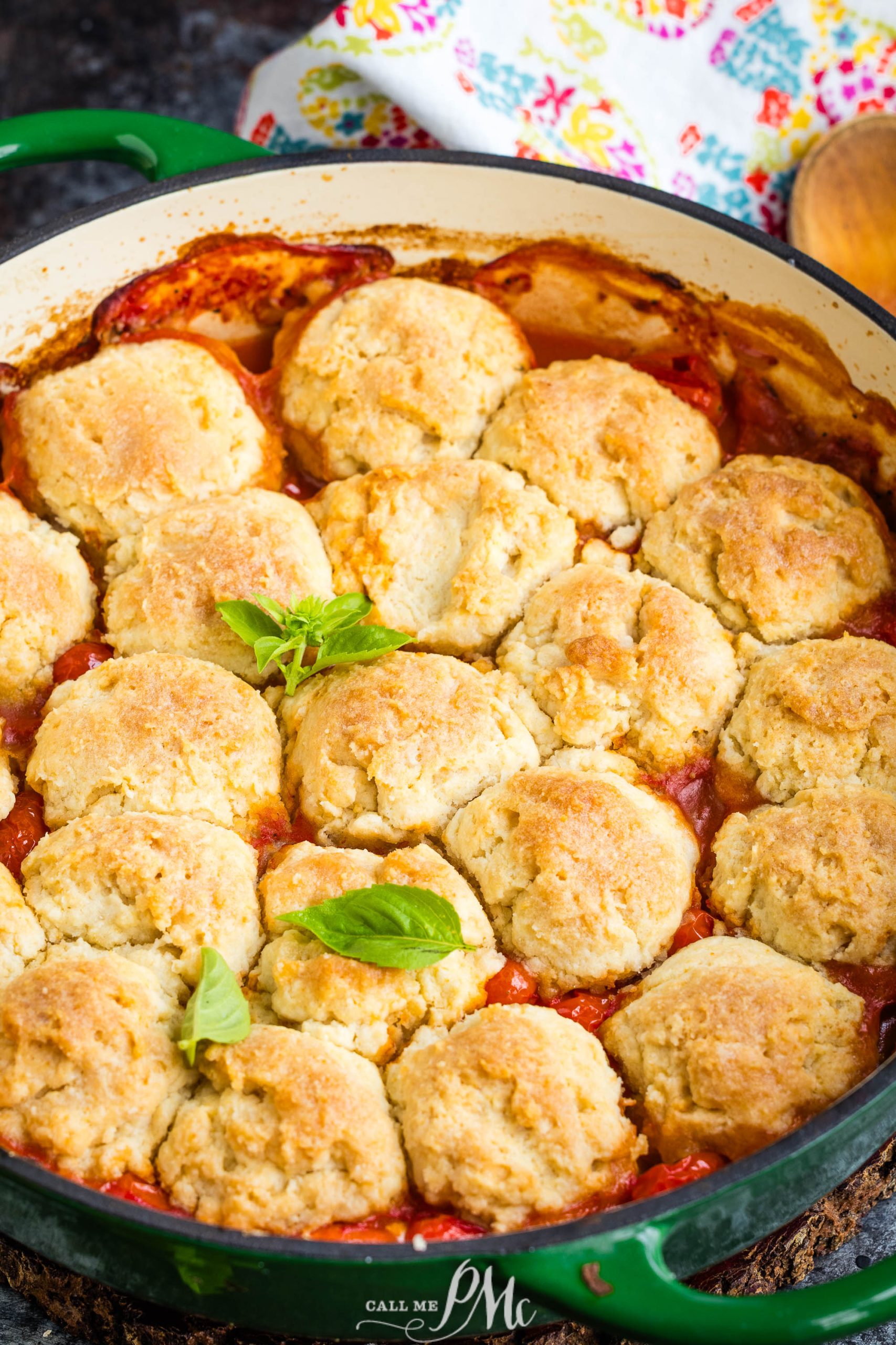 A round baking dish filled with golden-brown biscuits on top of a tomato-based casserole, garnished with fresh basil leaves. A colorful cloth is partially visible in the background.