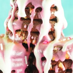 A stack of chocolate waffles drizzled with pink and white icing, topped with sprinkles and decorative pearls, against a light blue background.