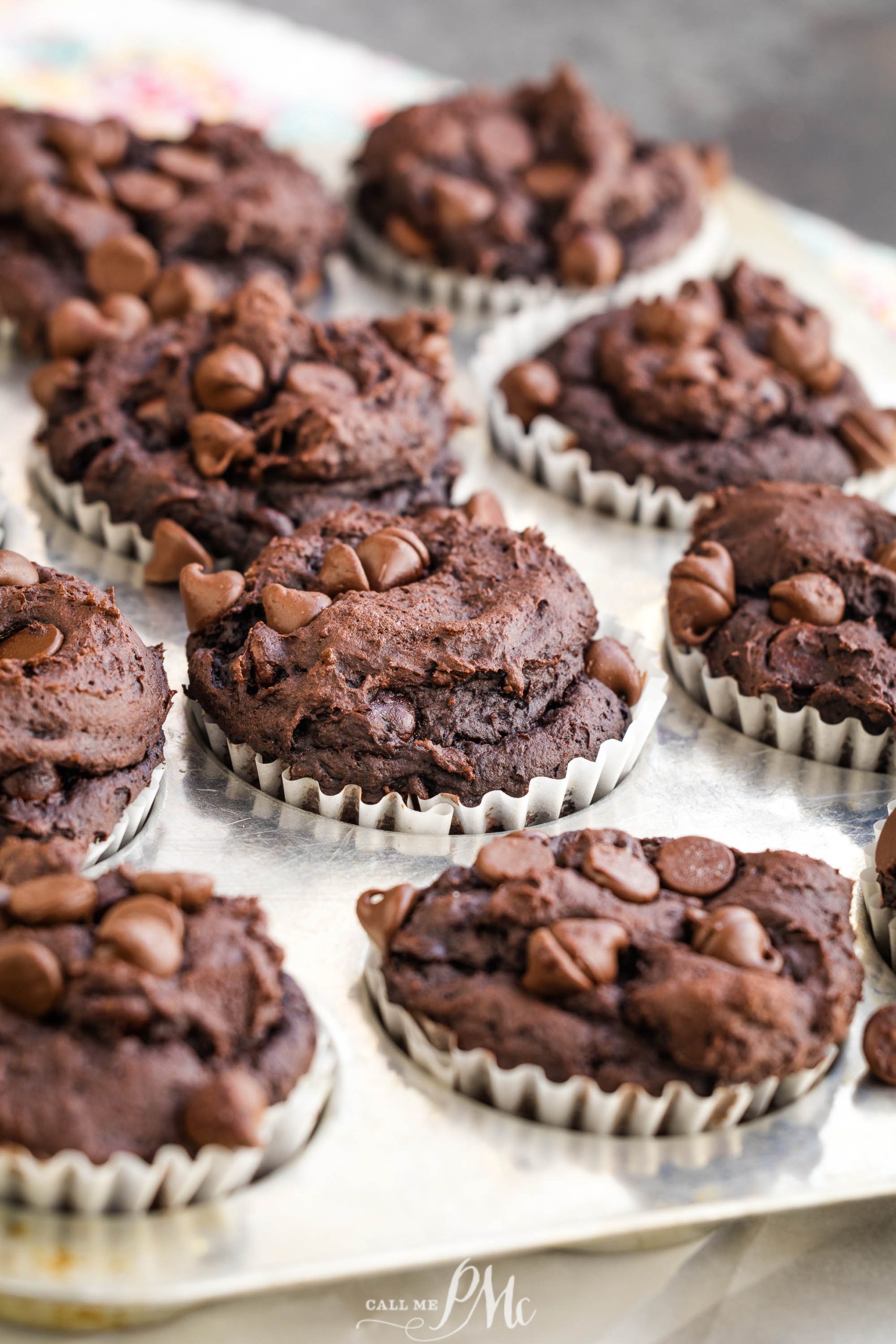 A batch of chocolate muffins topped with chocolate chips, placed in white paper liners on a baking tray.